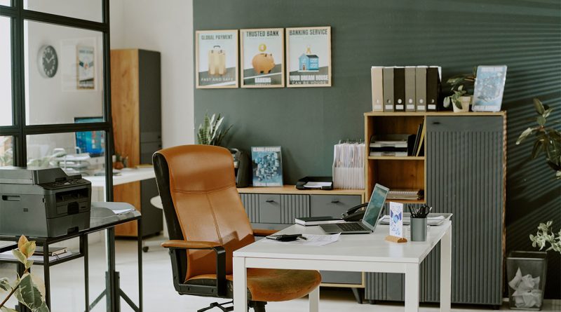Modern office space with a brown leather chair, white desk, laptop, printer, and organized shelves with files and folders against a green wall.
