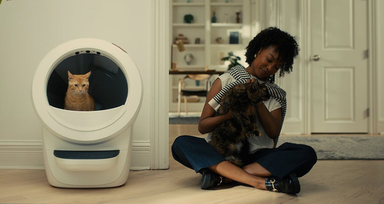 a woman sitting on the floor of a modern room, petting a dark-furred cat. To her left, an orange cat is visible inside a large, white, self-cleaning litter box, which is identified as a Litter-Robot 4 by Whisker.