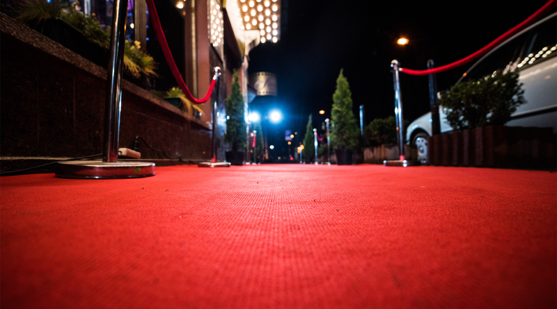 Low-angle view of a red carpet walkway at night, lined with velvet ropes and lights outside a venue entrance.